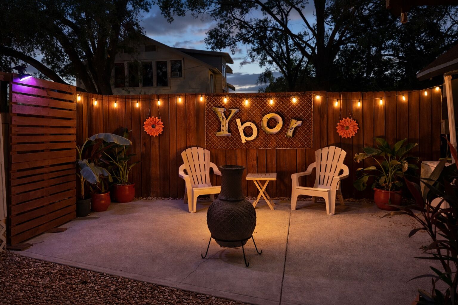 A patio with two Adirondack chairs, a table, a decorative vase, and a wooden fence with string lights.