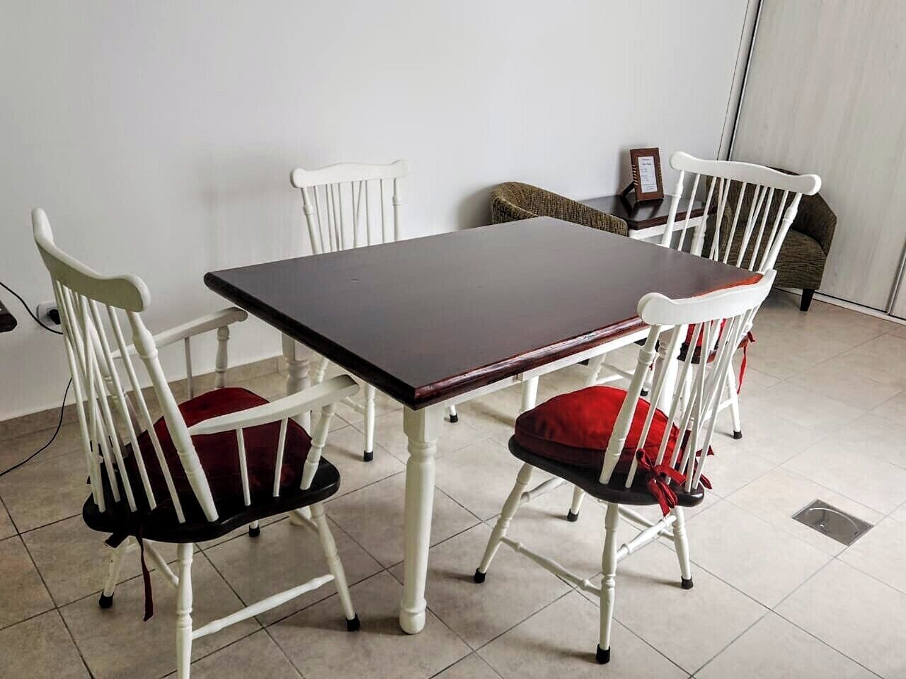 Uma sala de jantar com uma mesa retangular e quatro cadeiras brancas com almofadas vermelhas.