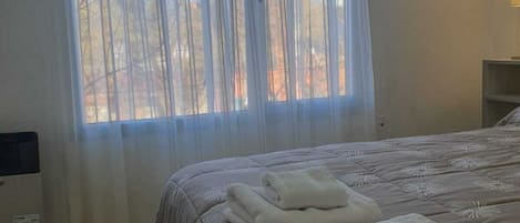 A bedroom with a bed, a window with sheer curtains, and a bedside table with a lamp.