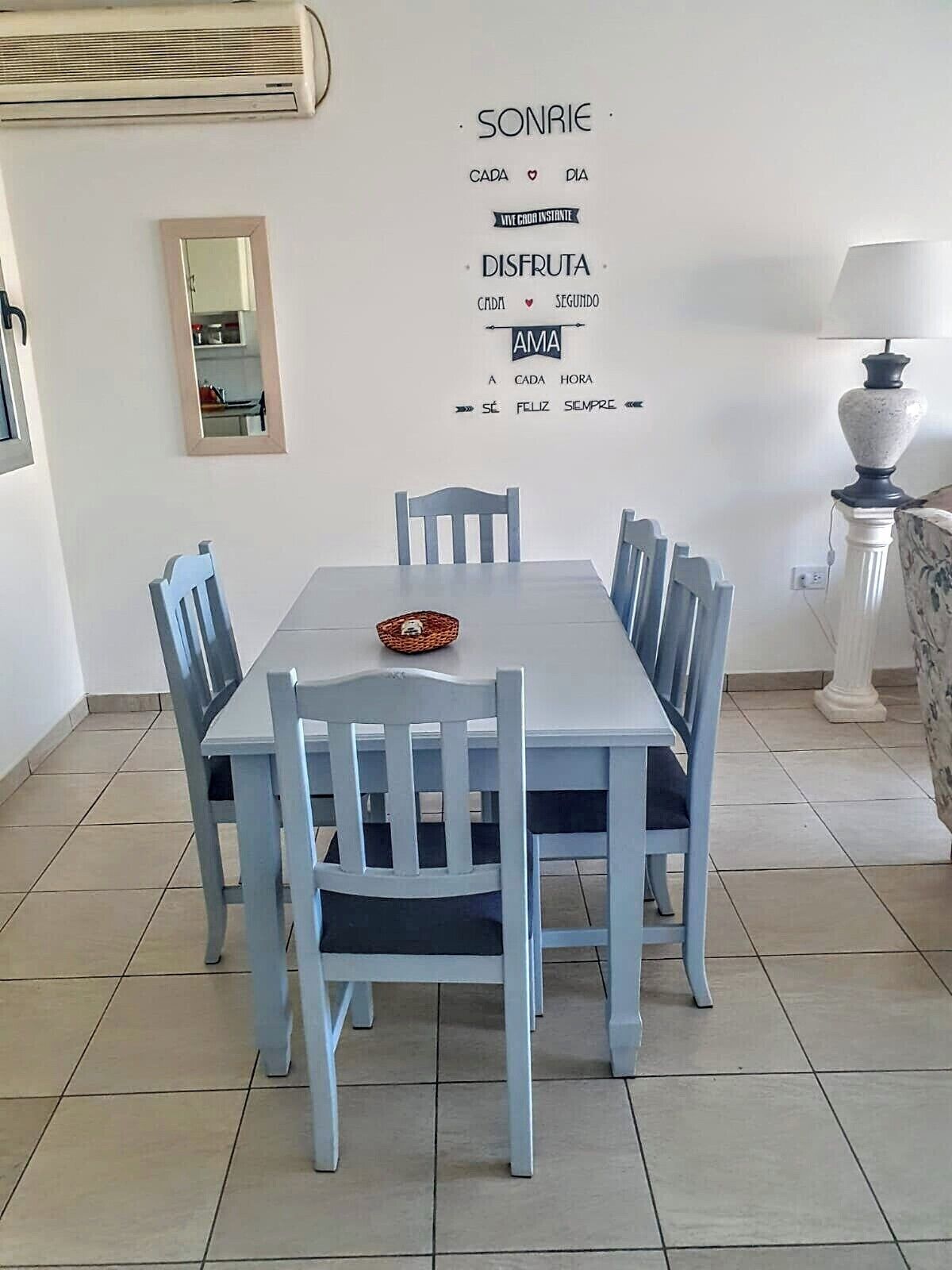 Un comedor con una mesa blanca y sillas azules, un espejo montado en la pared y un cartel decorativo con mensajes positivos.