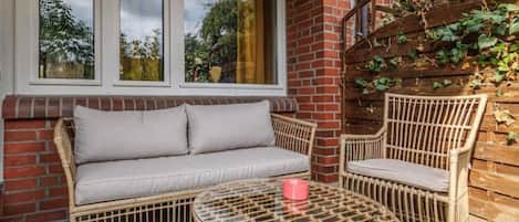 A wicker sofa and chairs near a window with a view of trees, a brick wall, and a wooden trellis with ivy.