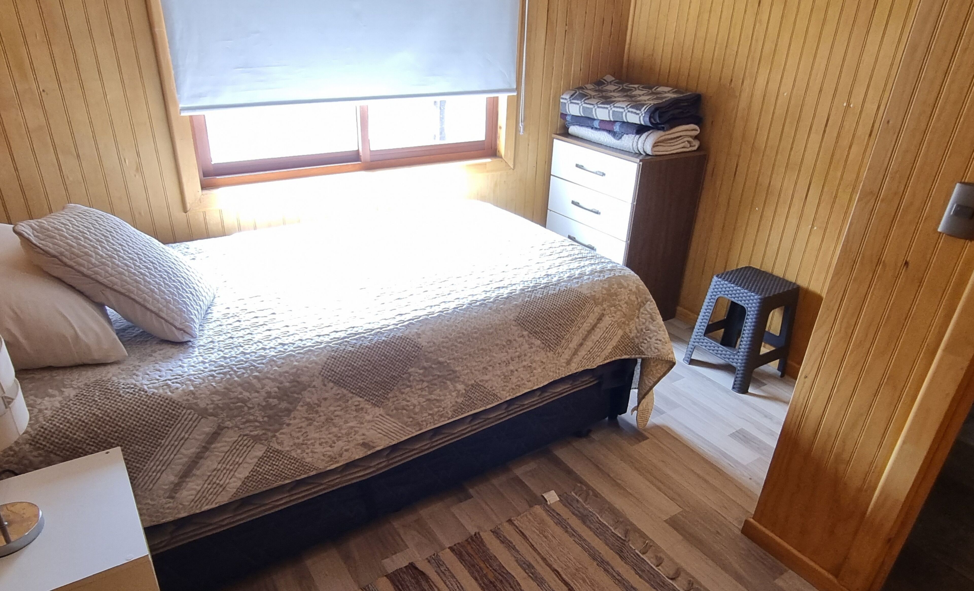 A small, cozy bedroom with a bed, a nightstand, a window with blinds, and a wooden wardrobe.