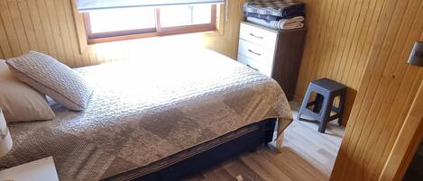 A small, cozy bedroom with a bed, a nightstand, a window with blinds, and a wooden wardrobe.