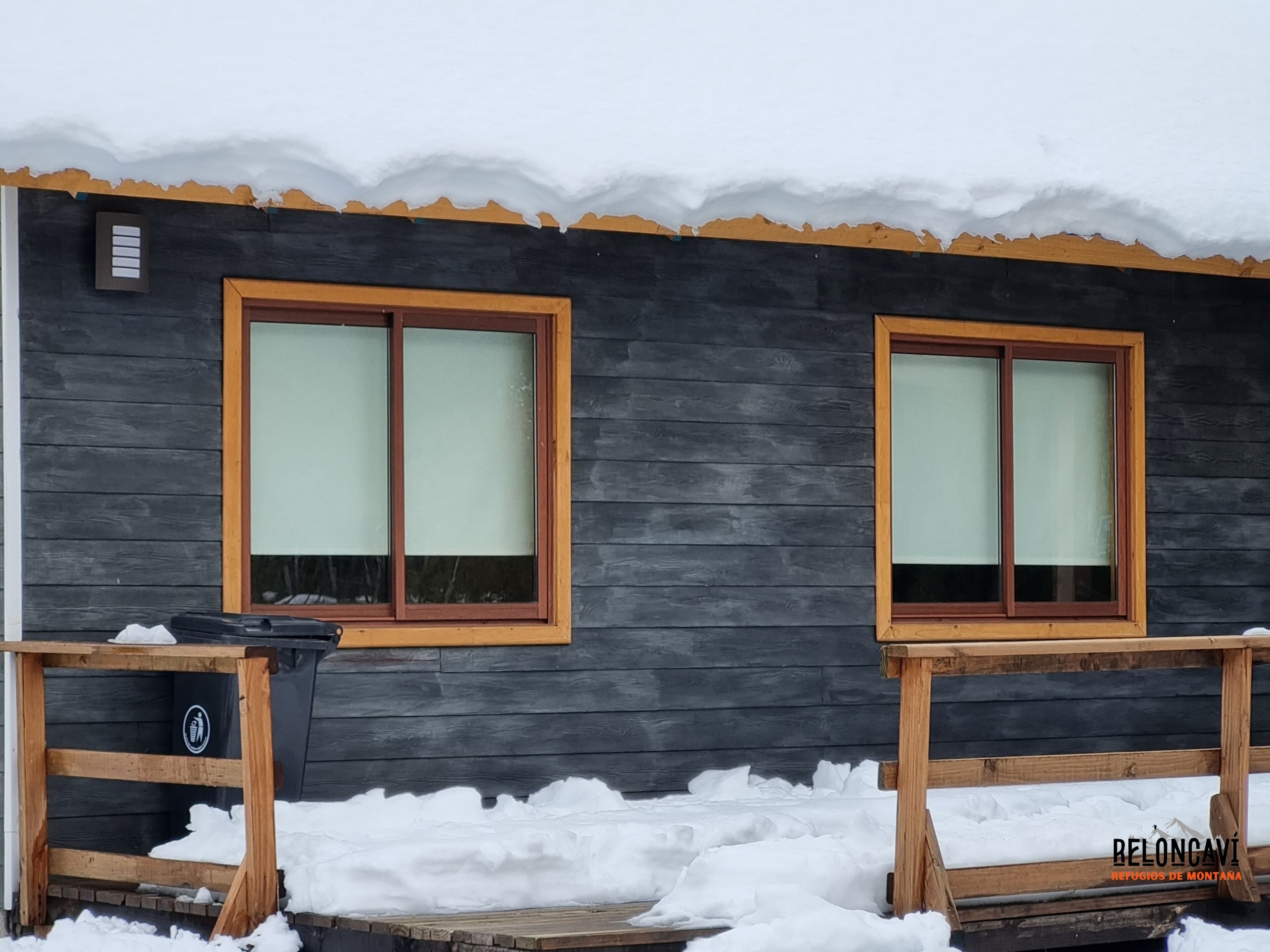 Eine Holzhütte mit zwei großen Fenstern, einer Holzterrasse und einer schwarzen Außenwand.