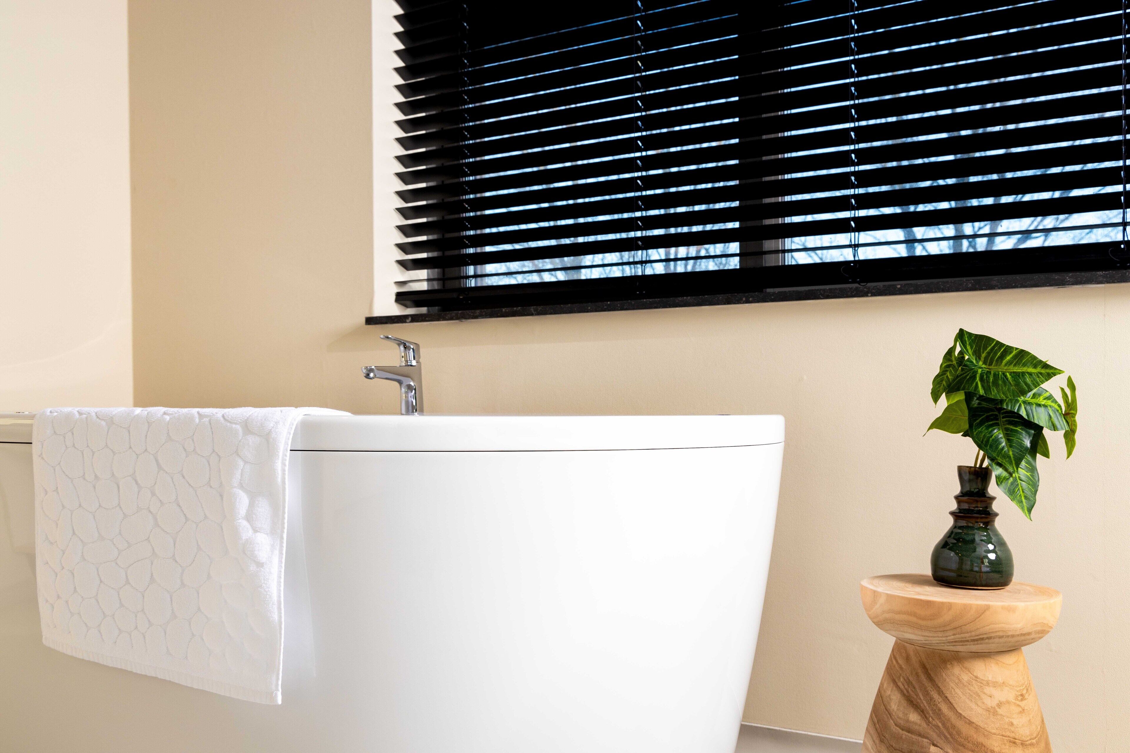 Un baño con una bañera blanca, una toalla colgada en la pared, un taburete de madera con una planta y persianas negras en la ventana.