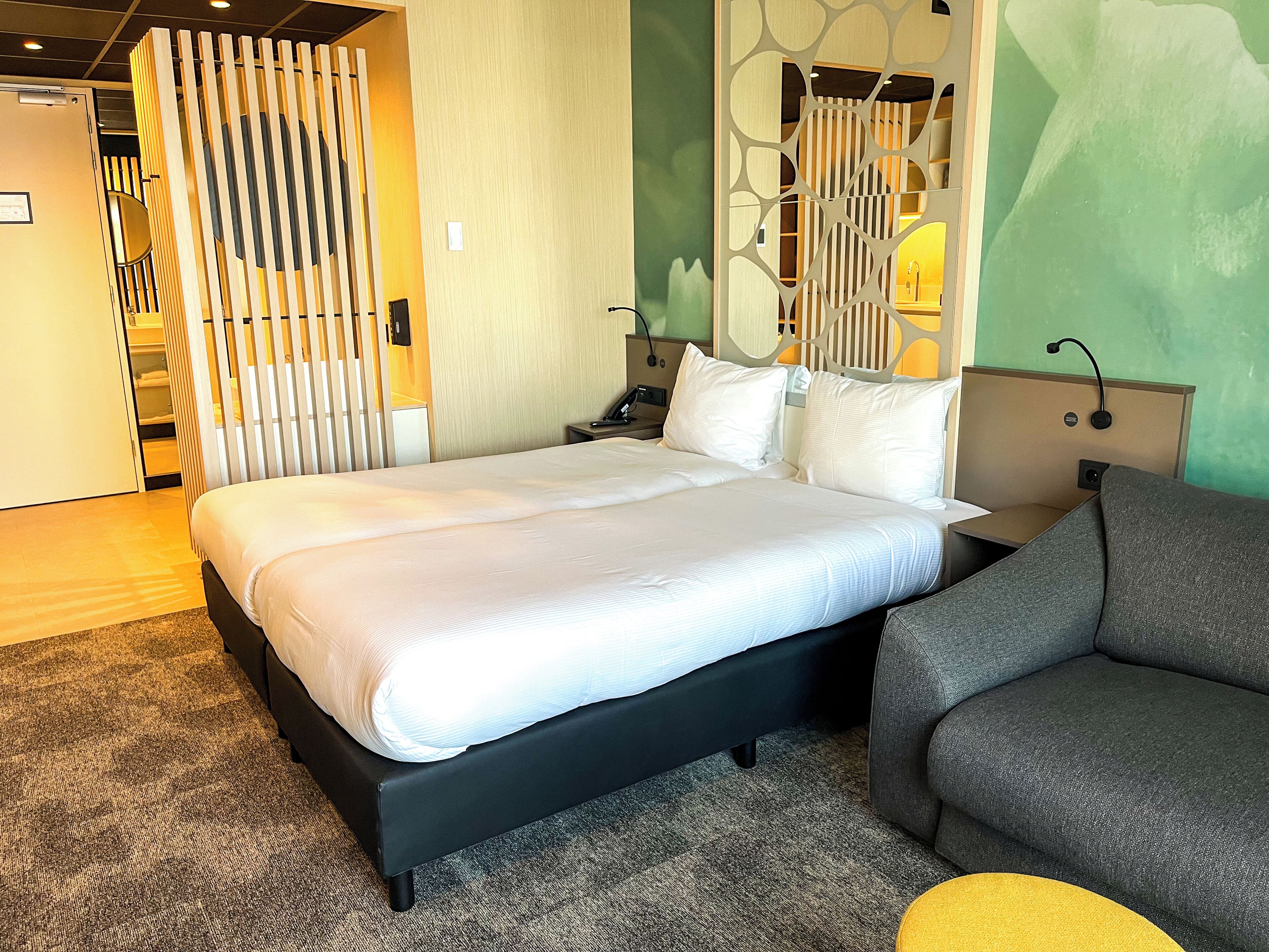 A hotel room with a large bed, a desk, a chair, and a partition with vertical slats.