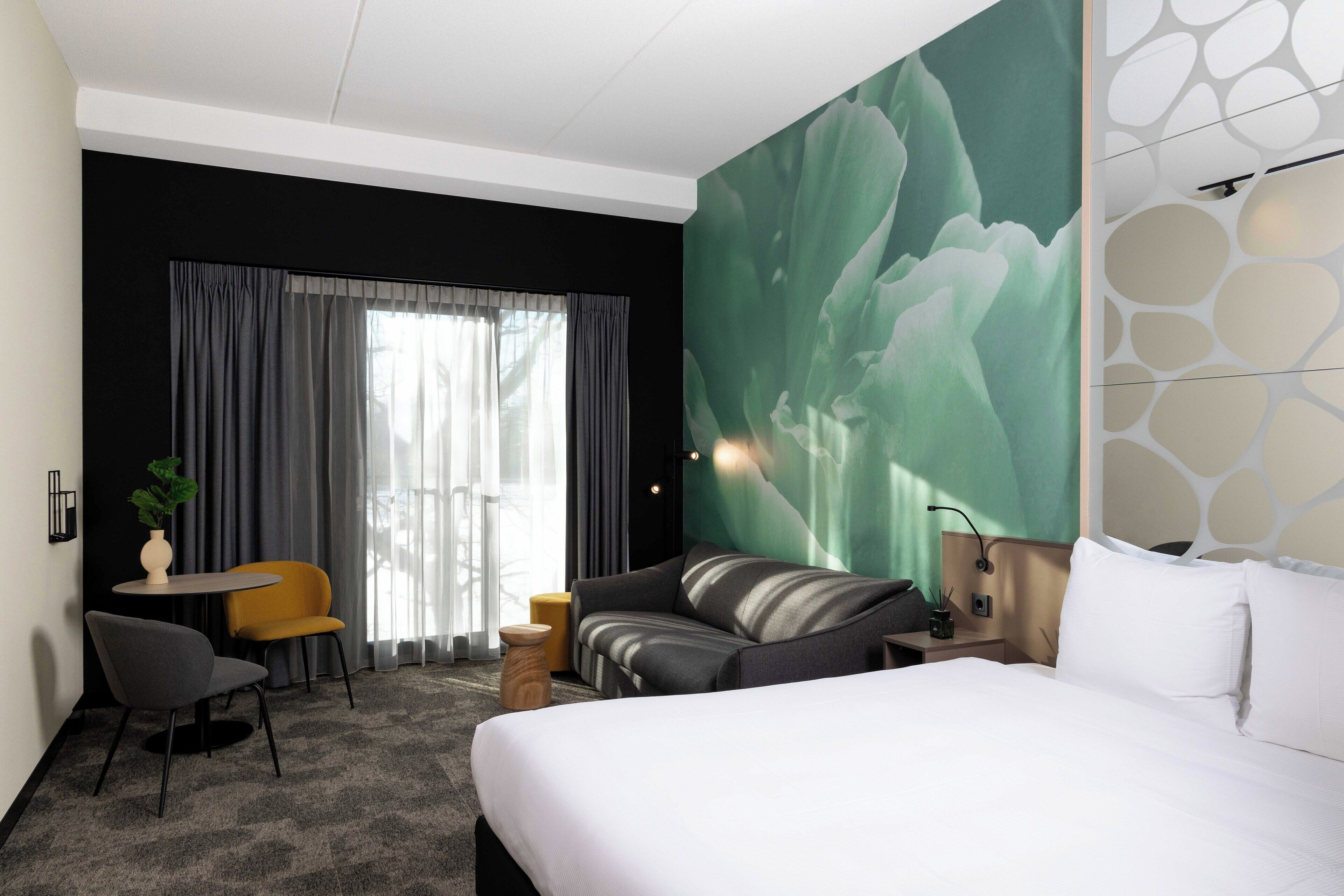 A hotel room with a large bed, a sofa, a small table with a plant, and a view of a cityscape through the window.