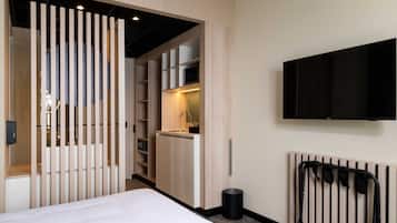 Habitación de hotel con cama, televisión montada en la pared, armario empotrado y una pequeña cocina.