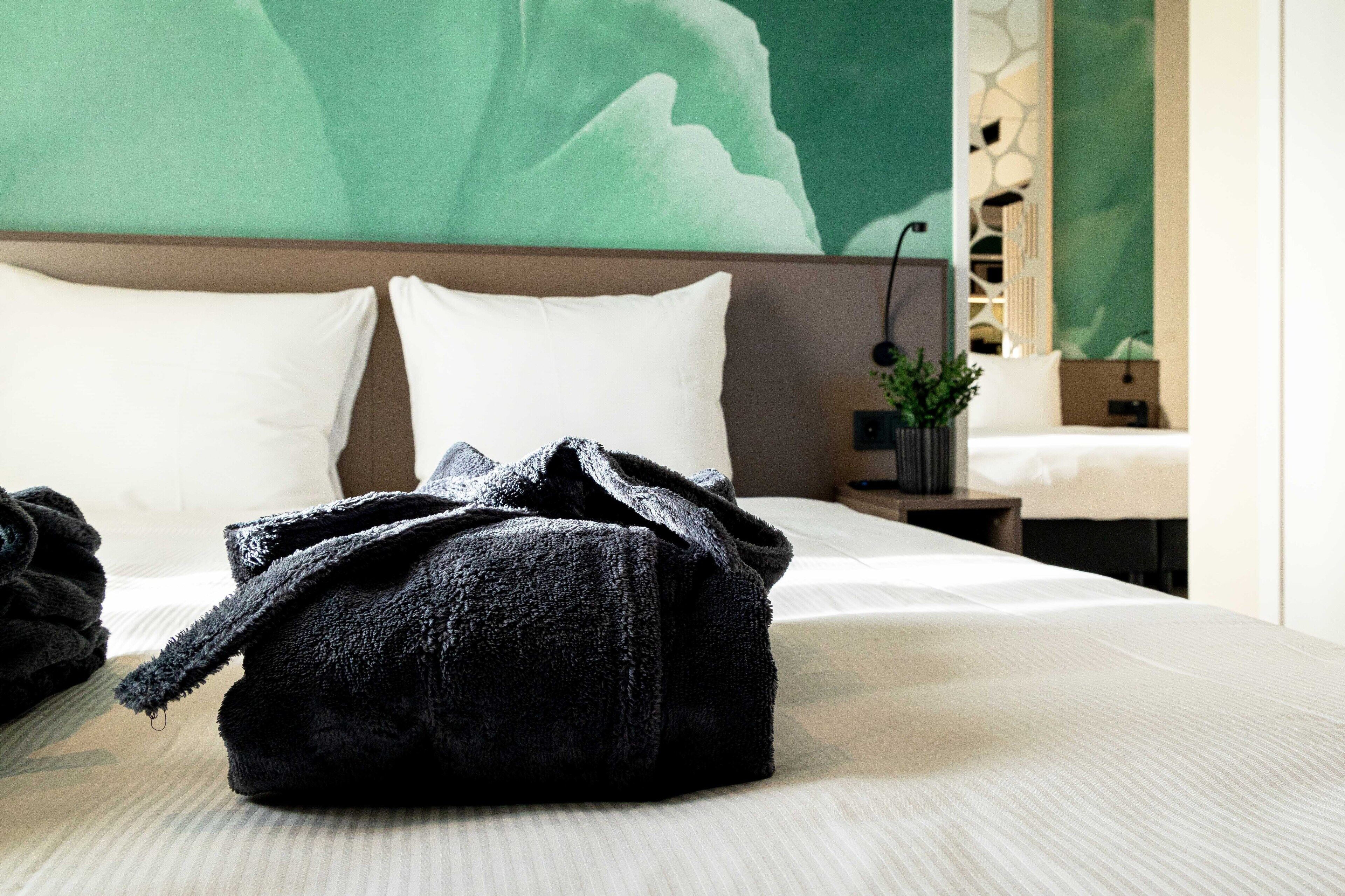 Una cama bien hecha con un bolso negro encima, una pared verde y una planta pequeña sobre una mesita de noche.