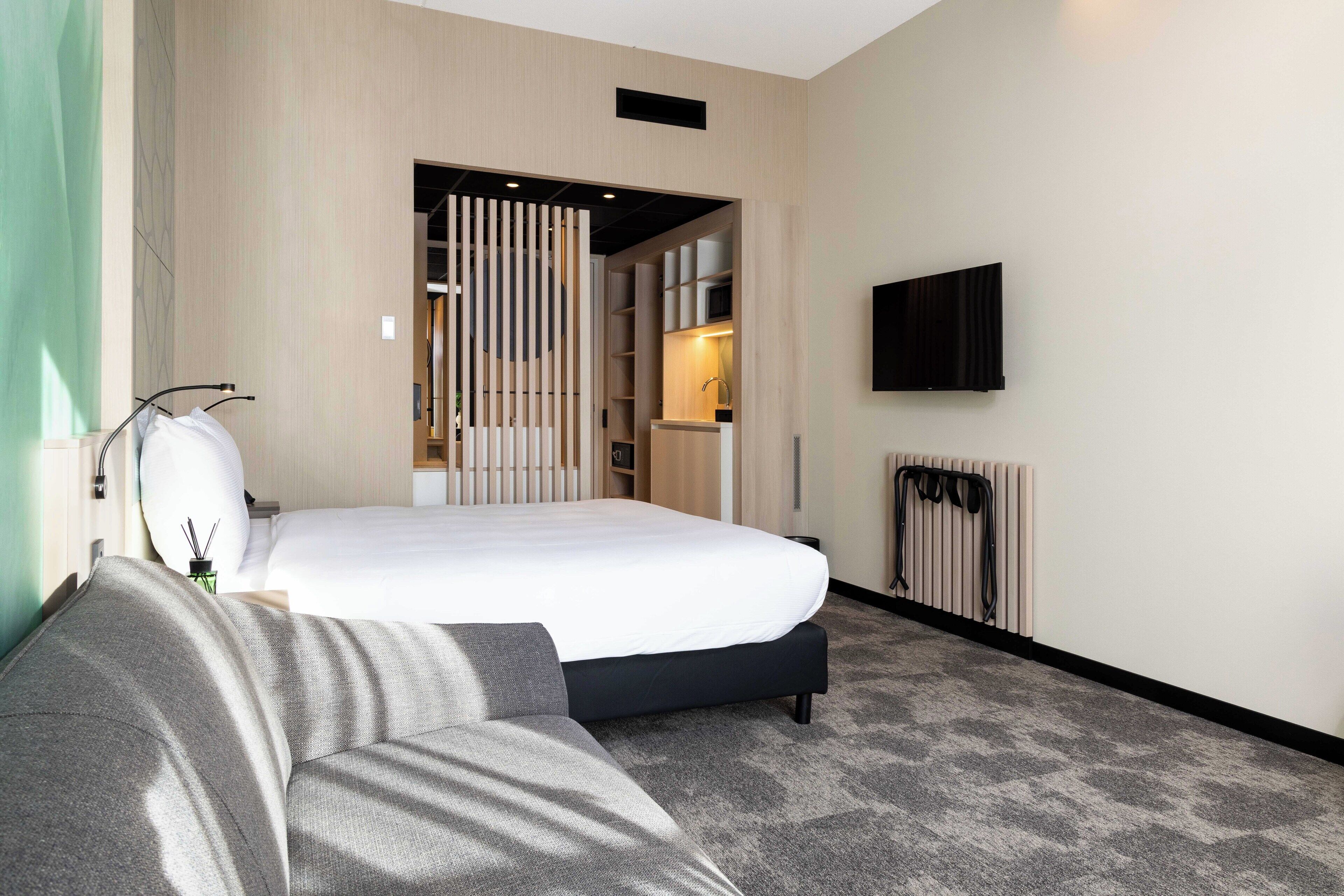 A modern hotel room with a large bed, a sofa, a TV mounted on the wall, and a radiator.