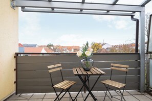 A balcony with a wooden table and two chairs, a vase with flowers, and a view of buildings in the background. - Homaris Apartments München Laim (Munich)
