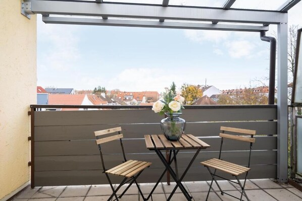 A balcony with a table and two chairs, a vase with flowers, and a view of houses in the background. - Homaris Apartments München Laim (Munich)