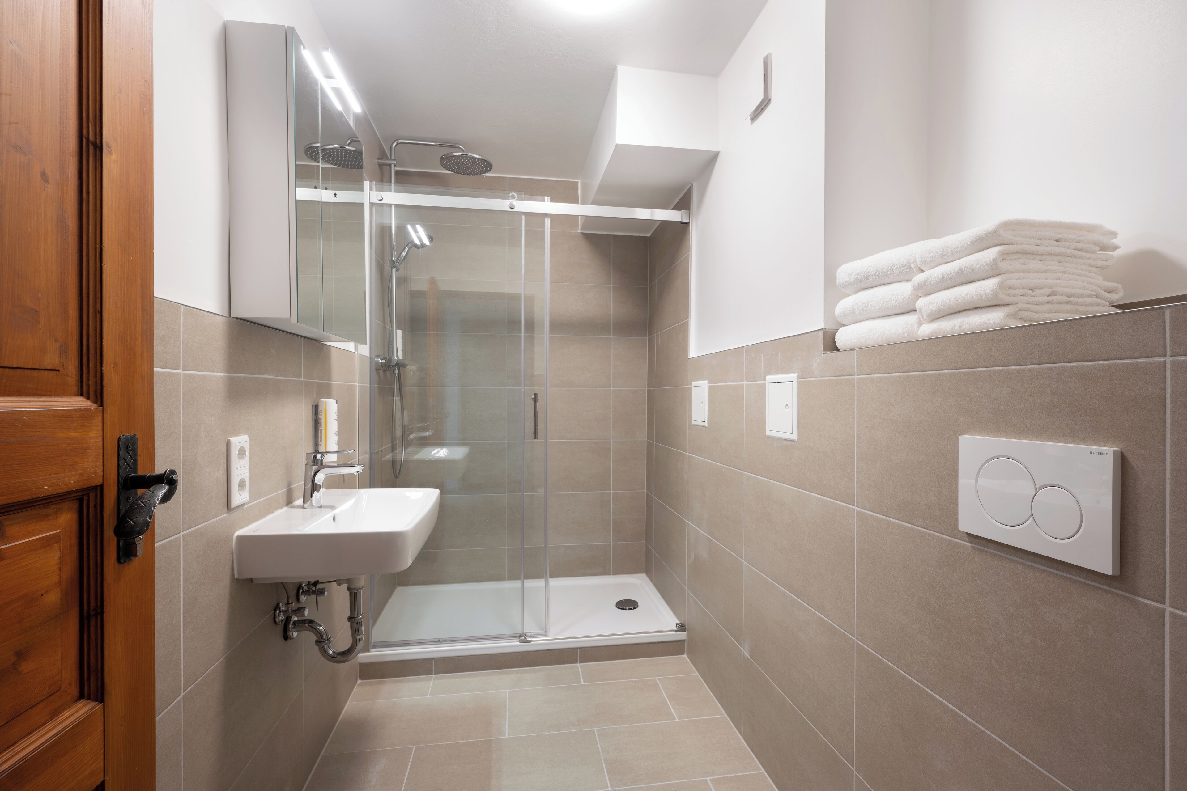 Un baño moderno con ducha de vidrio, lavamanos y una pila de toallas blancas en la pared.