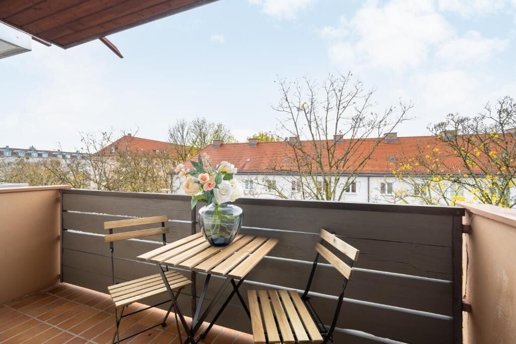 A balcony with a table and chairs, a vase of flowers, and a view of buildings and trees.