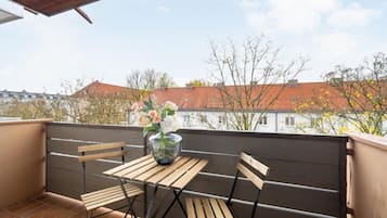 A balcony with a table and chairs, a vase of flowers, and a view of buildings and trees.