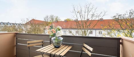 A balcony with a table and chairs, a vase of flowers, and a view of buildings and trees.