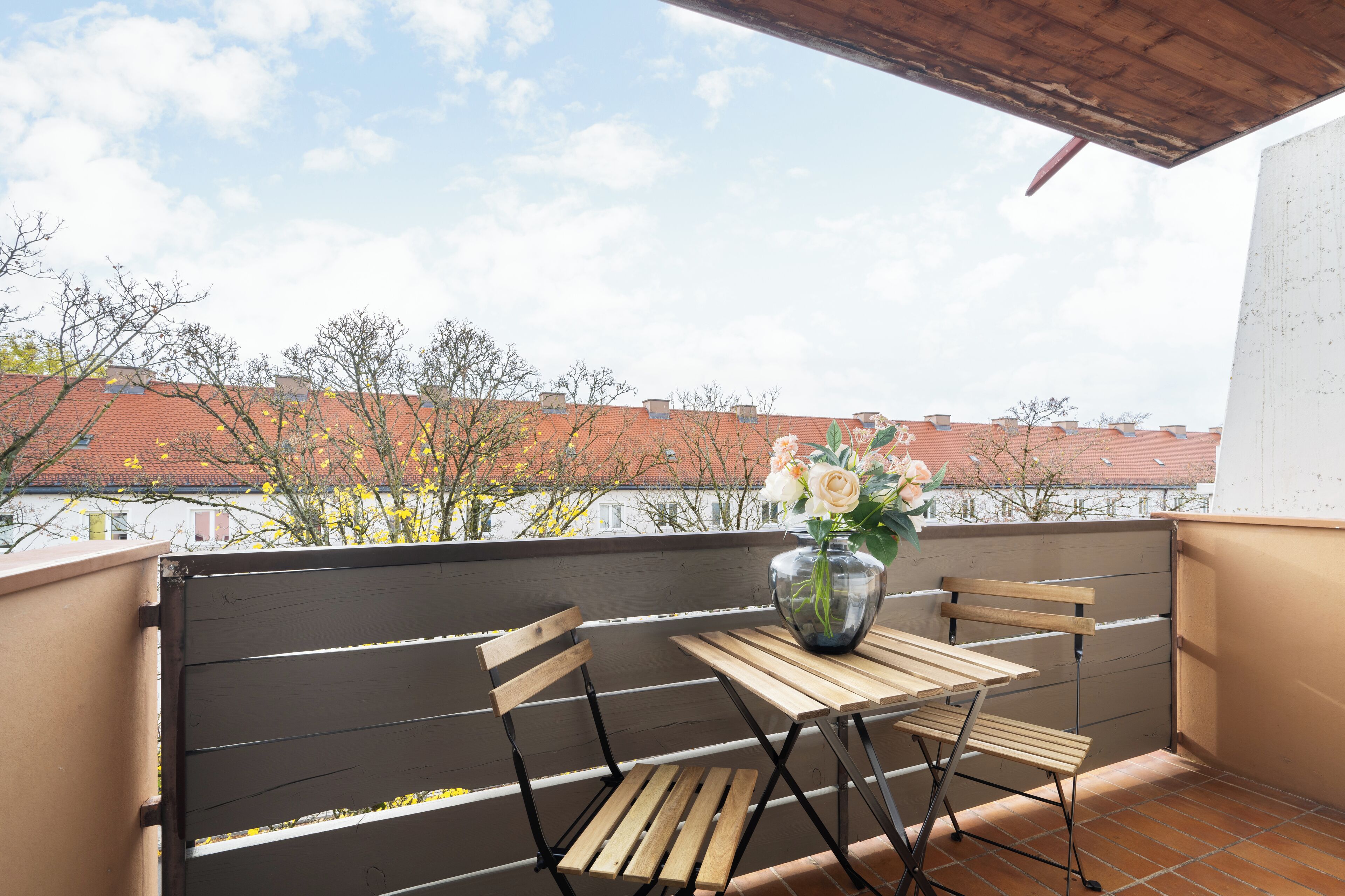 Un balcón con mesa y sillas de madera, un florero con flores y vistas a edificios y árboles.