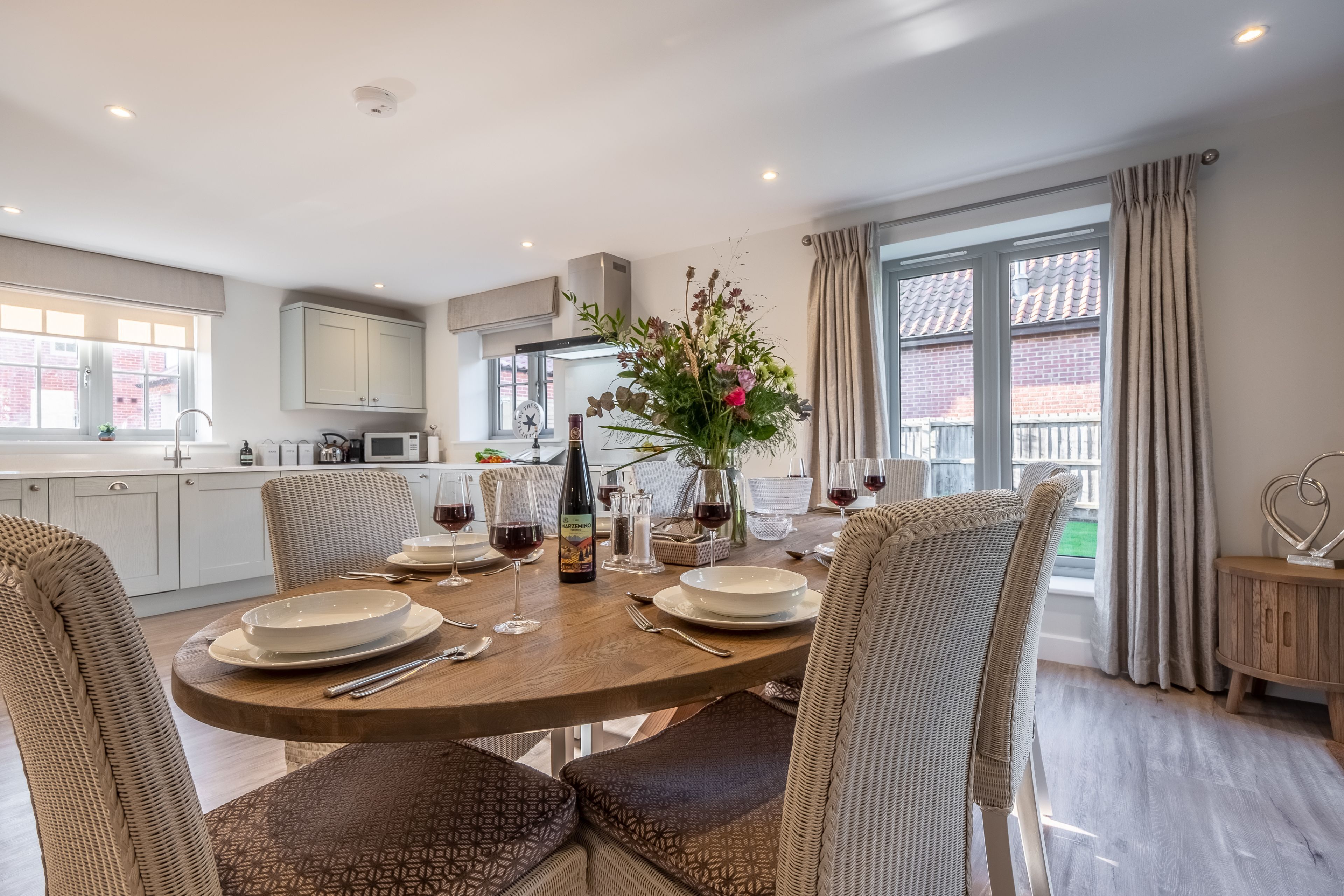 Een eethoek met een ronde tafel gedekt voor vier personen, omringd door rieten stoelen. Op de tafel staat een vaas met bloemen, en een fles wijn is zichtbaar. Achtergrond toont een moderne keuken met witte kastjes en een raam met gordijnen.