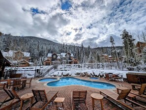 Un área de piscina con sillones reclinables y una terraza de madera, rodeada de árboles nevados y edificios.