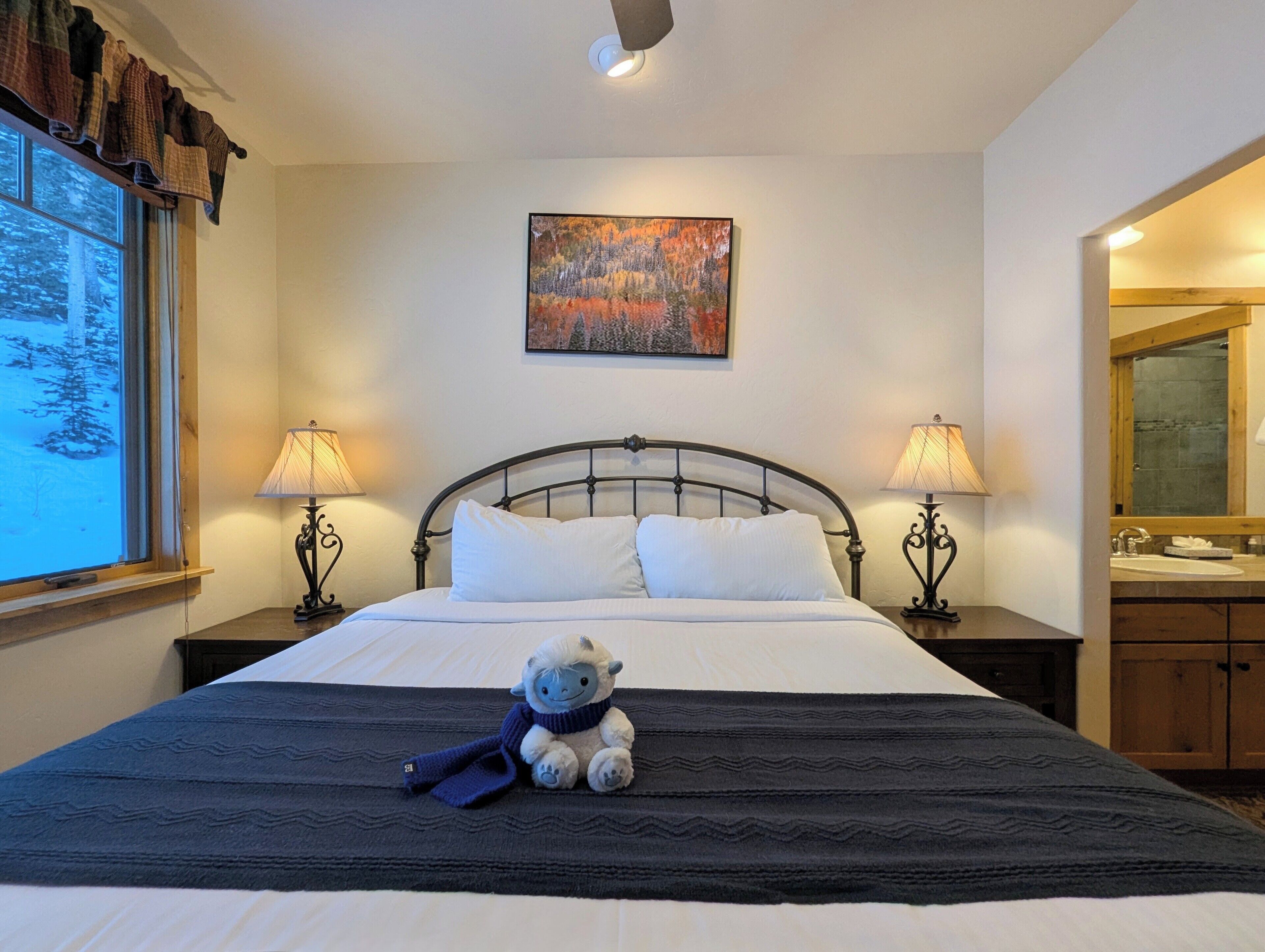 A neatly made bed with white linens, two bedside lamps, a painting above the bed, and a window with a view of trees.