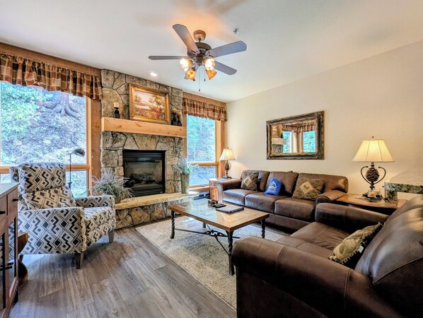 A living room with a stone fireplace, a ceiling fan, a sofa, a patterned armchair, and a coffee table. - Stylish Lodge with Pool/Hot Tub/Shuttle (Keystone)