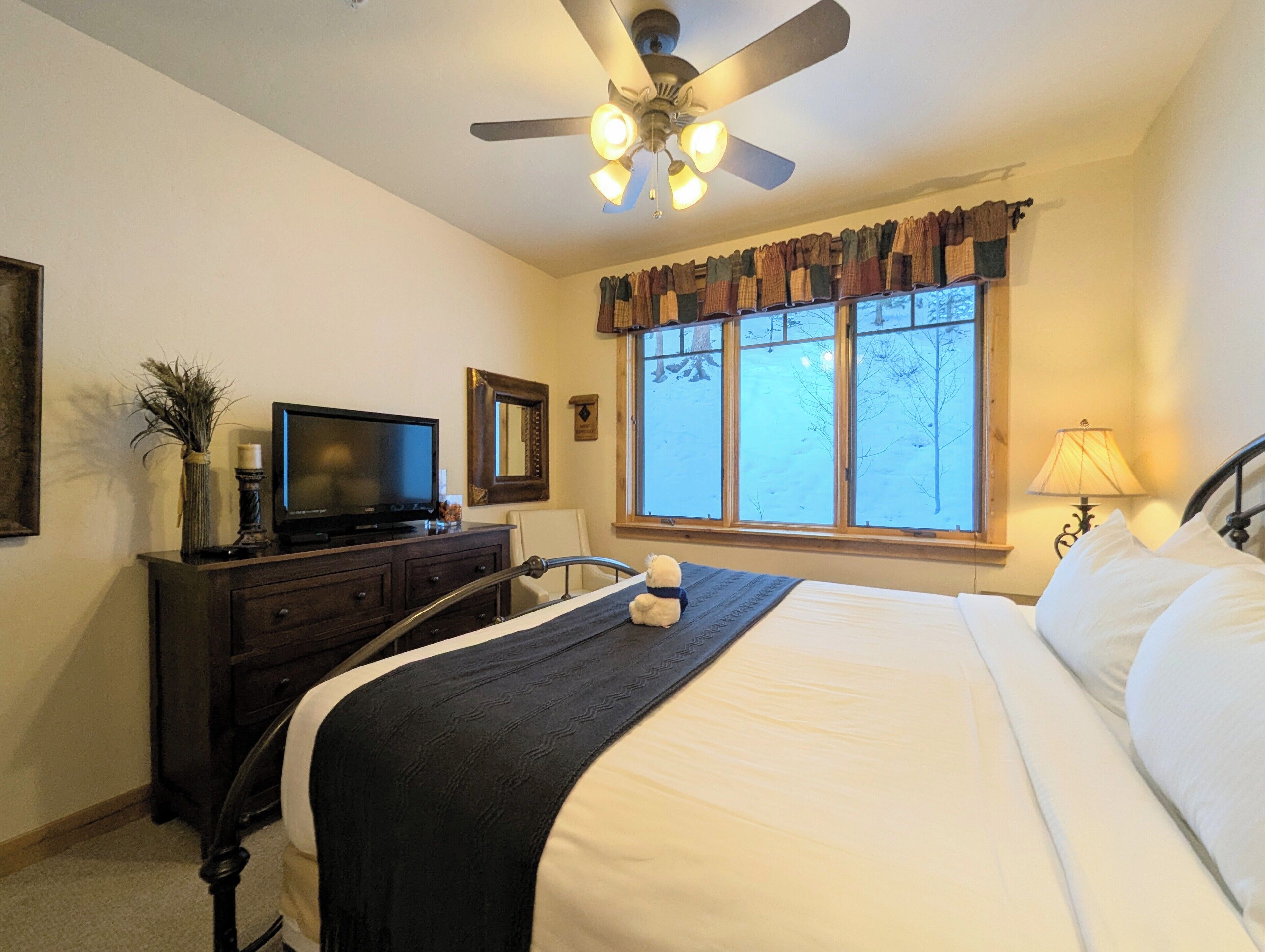 A bedroom with a bed, a television on a dresser, a window with curtains, a ceiling fan, and a lamp.