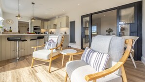 A modern kitchen with bar stools, a dining area with a round table, and a living room with a sofa and chairs. - Larks Rise, Burnham Market, Norfolk (Burnham Market)