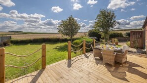 A wooden deck with wicker furniture and a table set for dining, overlooking a grassy field and a brick building. - Larks Rise, Burnham Market, Norfolk (Burnham Market)