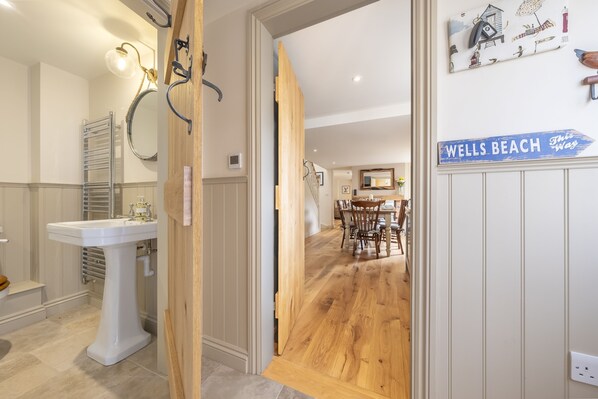 Ein Badezimmer mit einem weißen Waschtisch, einem Handtuchhalter und einem Spiegel. Eine Holztür führt in einen Essbereich mit Holzboden und einem Tisch mit Stühlen. Ein Schild an der Wand zeigt: "WELLS BEACH This Way".
