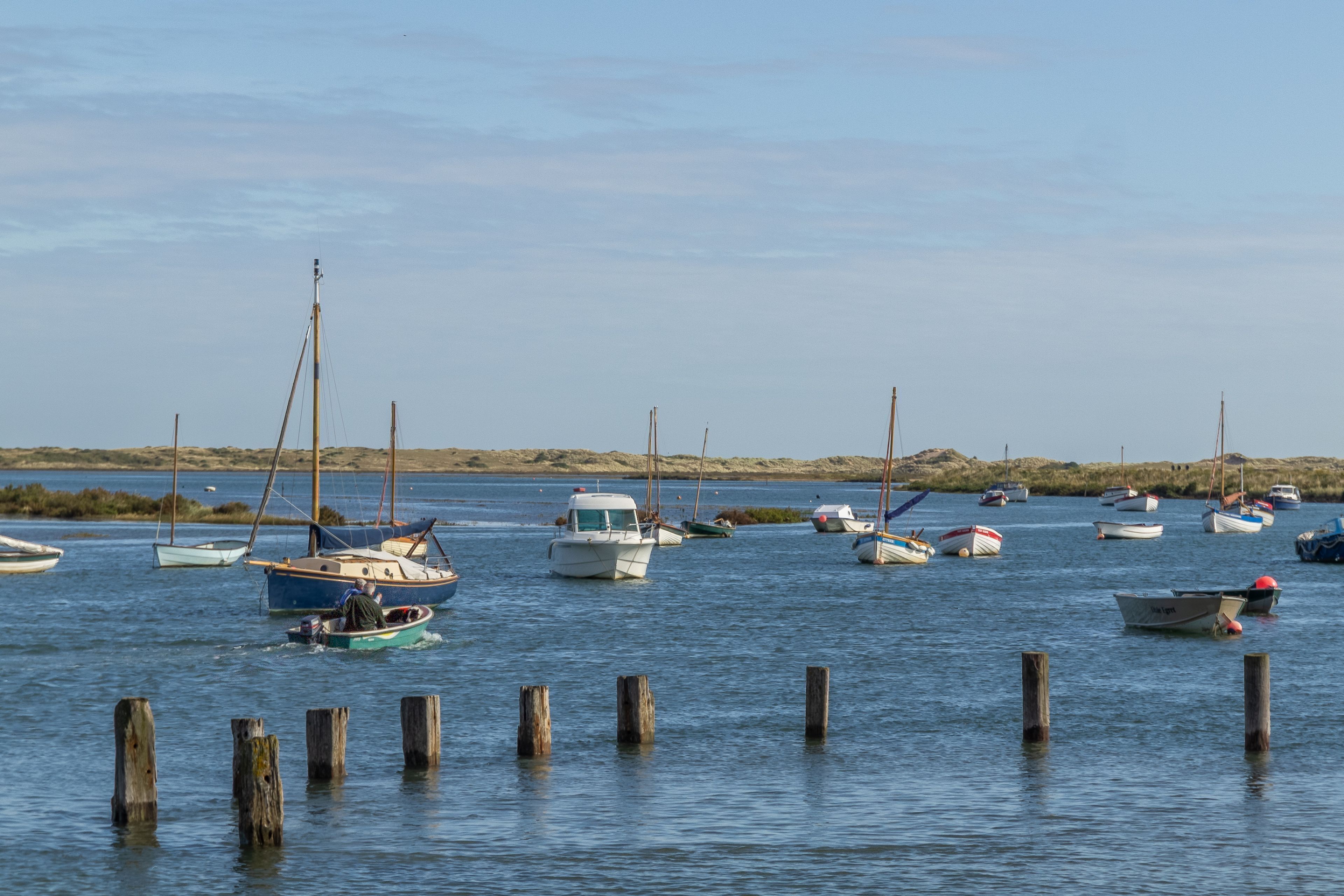 A marina with various boats docked, wooden posts in the water, and a sandy shoreline in the background.