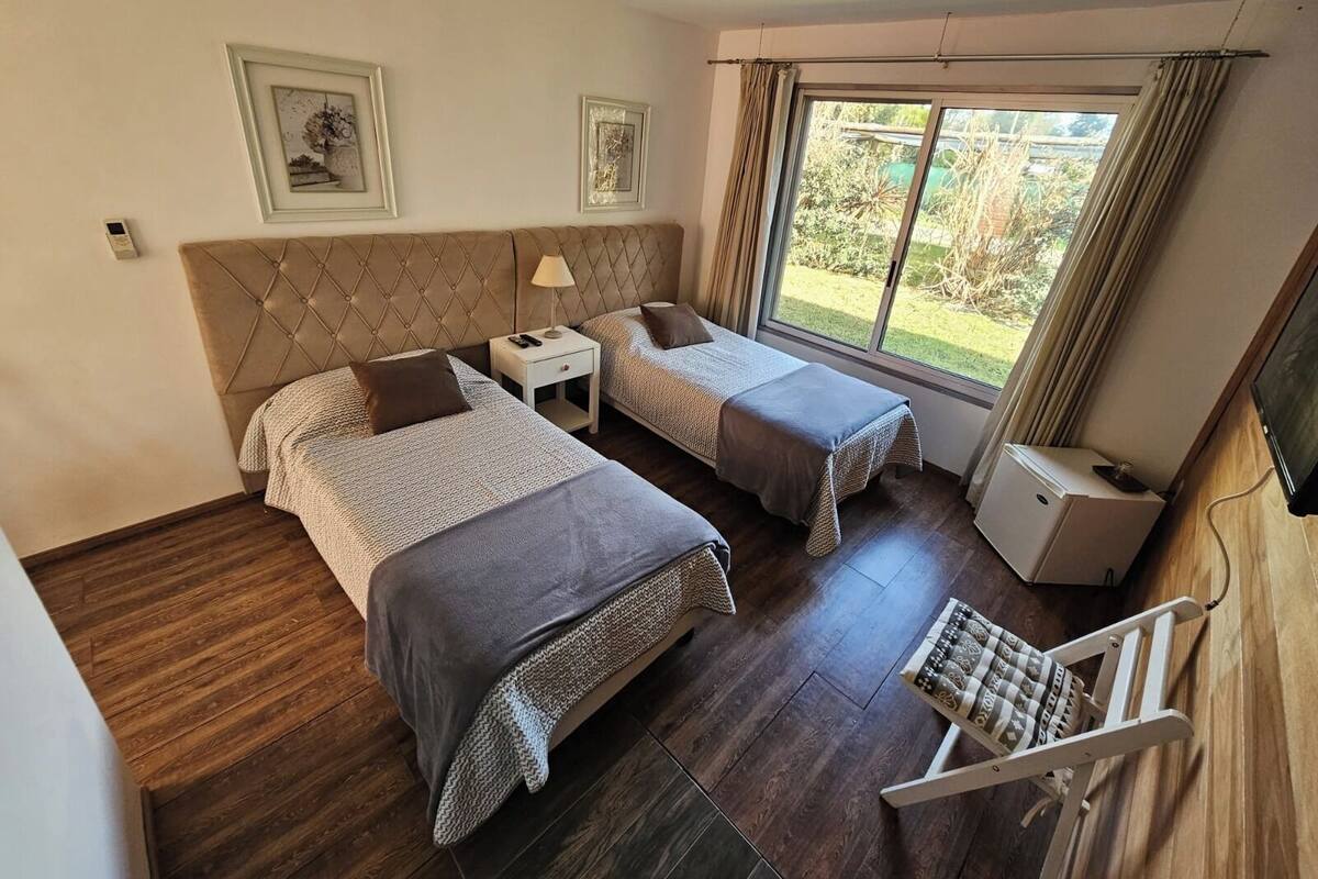 A room with two beds, a small table, a chair, and a window with a view of greenery.