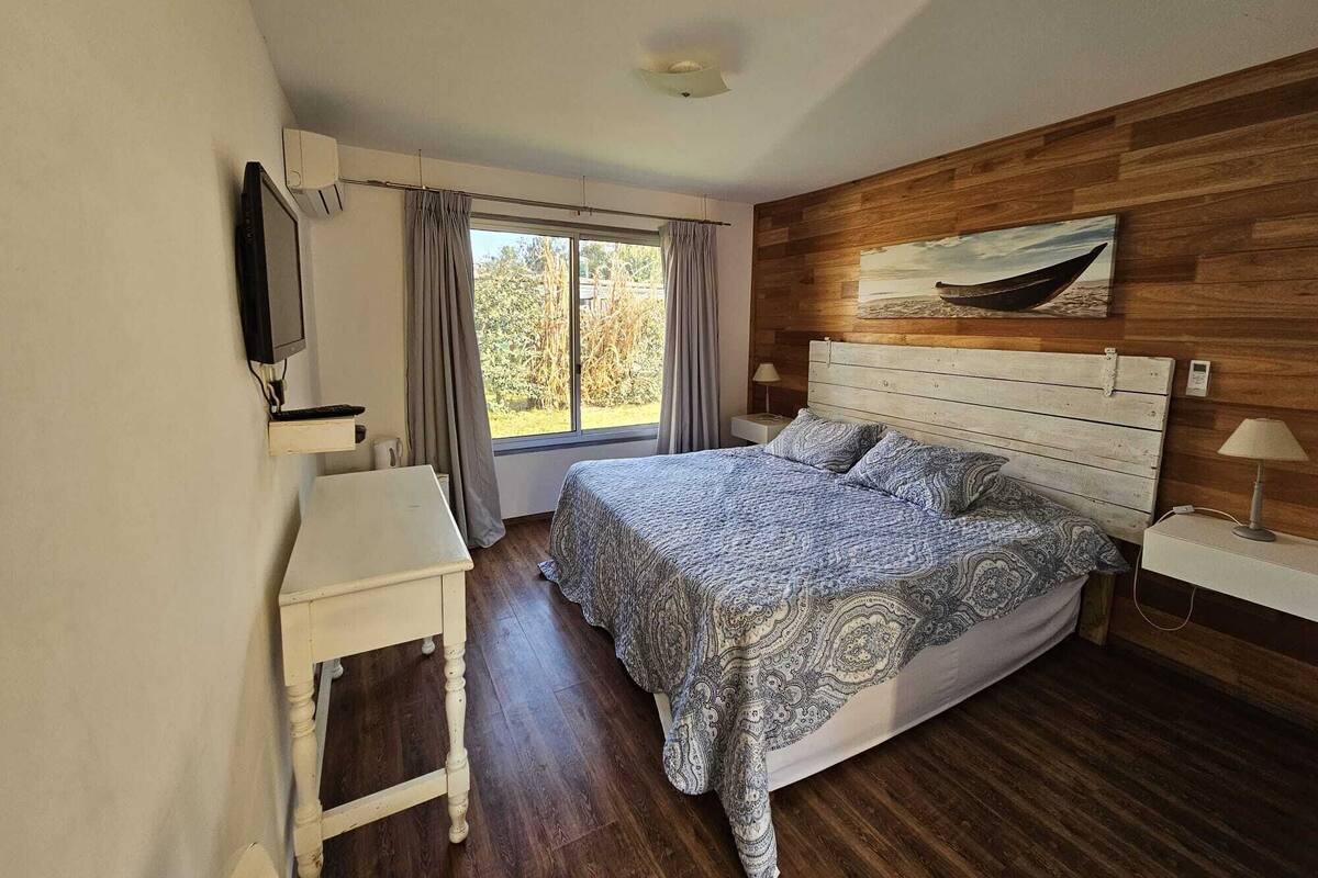A bedroom with a wooden headboard, a bed with patterned bedding, a bedside table, a TV mounted on the wall, and a window with curtains.