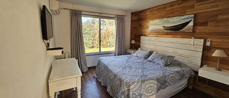 Un dormitorio con cabecera de madera, cama con ropa de cama estampada, mesita de noche, televisor montado en la pared y ventana con cortinas.