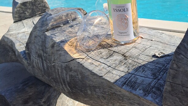 Une bouteille de vin Château Yssole sur une table en bois, près d’une piscine, avec une maison en arrière-plan.