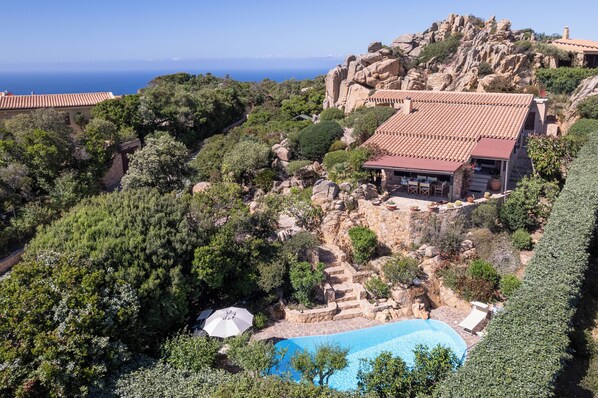 A villa with a swimming pool, surrounded by lush greenery and rocky terrain, overlooking a coastal area.