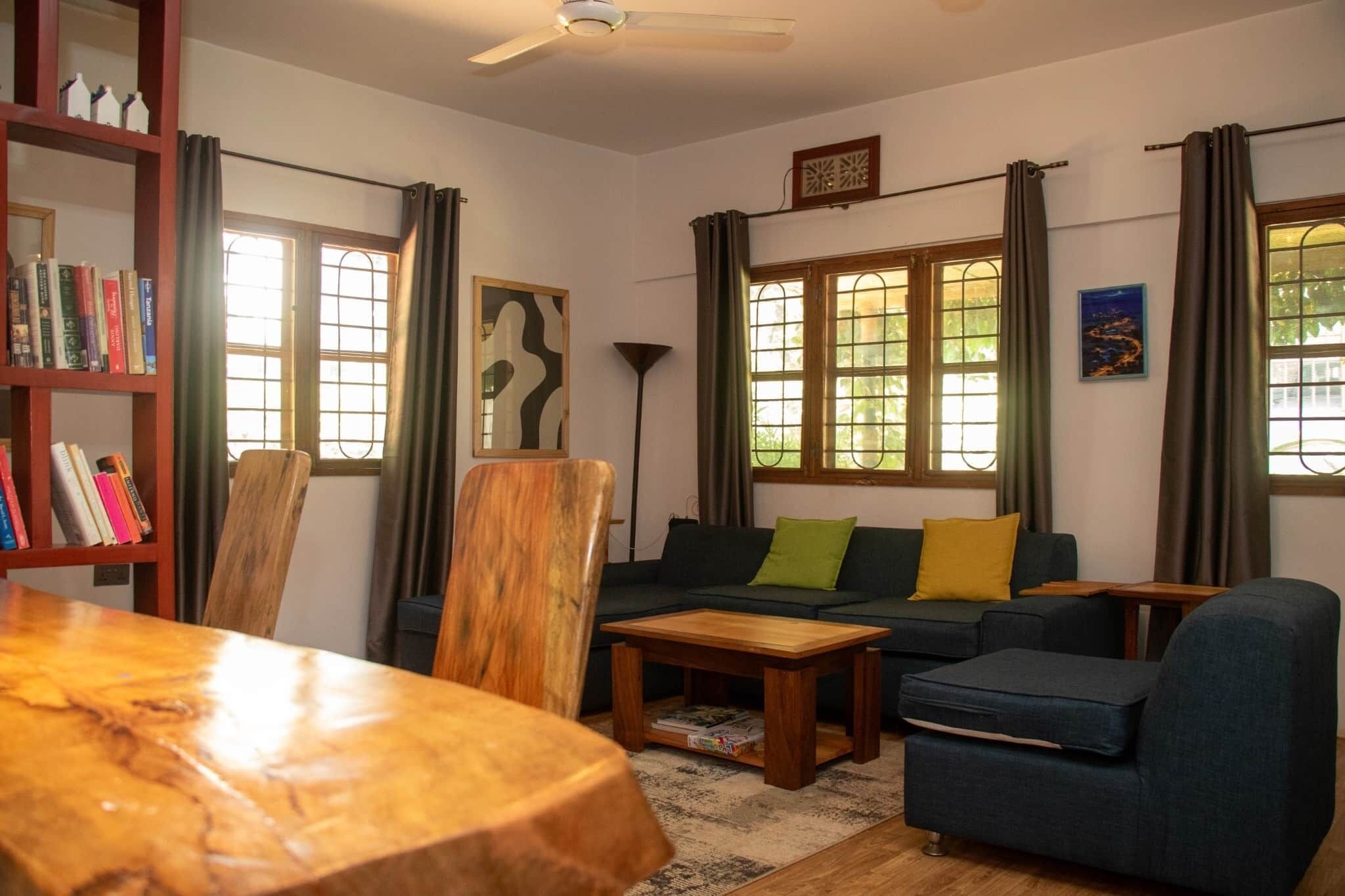 A living room with a wooden coffee table, a sofa with cushions, a bookshelf, and a large window with curtains.