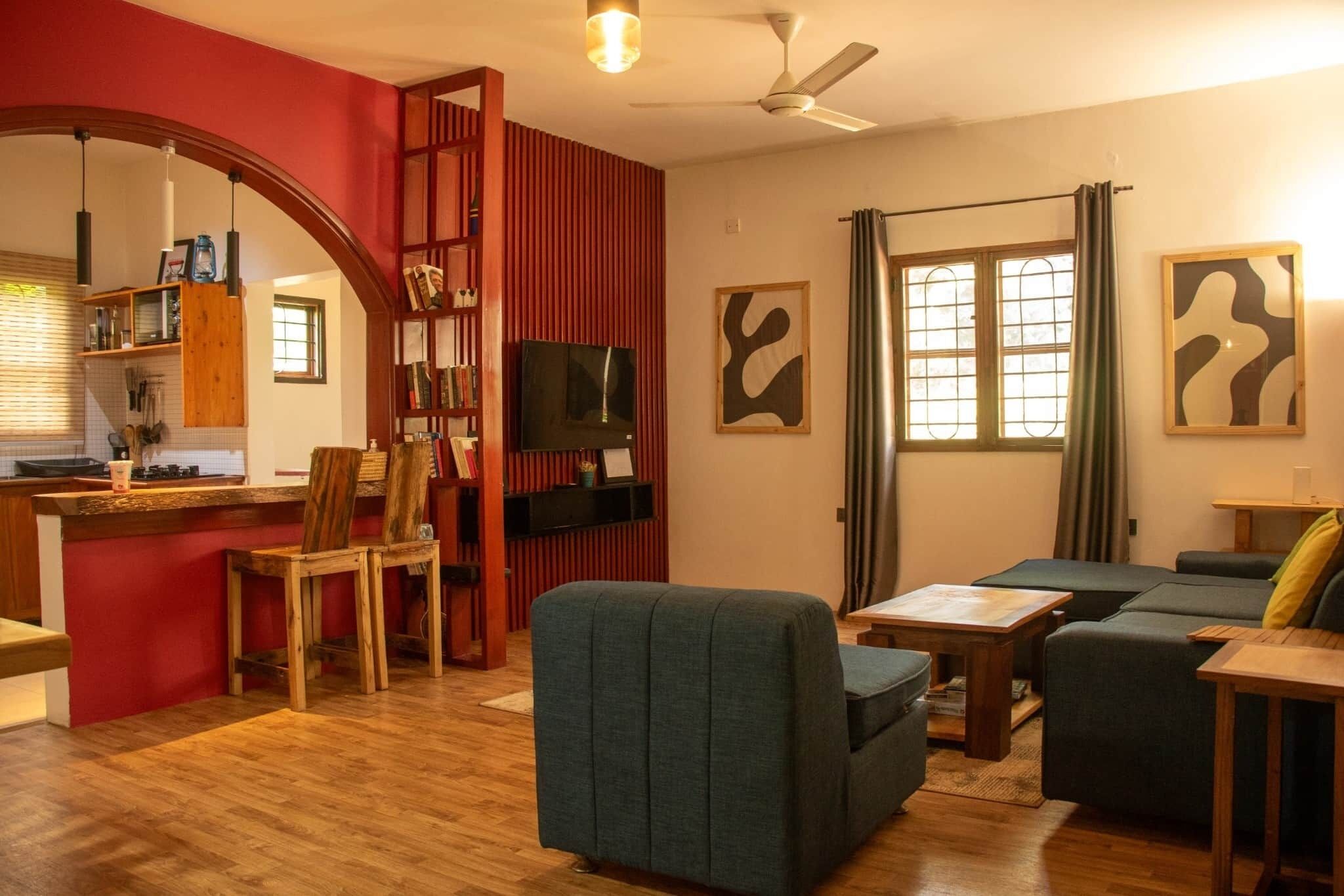 Una sala de estar con sofá, sillones, televisor y una zona de cocina con pared roja y estantes de madera.