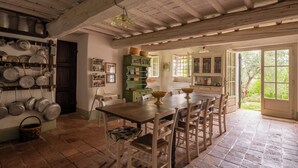 A rustic kitchen with a wooden table and chairs, a hanging pot rack, and a view of the garden through open doors.