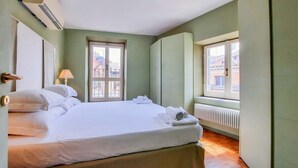 Un lit bien fait, avec du linge de lit et des serviettes blanches, une lampe et une fenêtre donnant sur des immeubles.