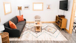 Un salon avec un canapé gris, des coussins orange, une table basse en bois, un petit fauteuil, un téléviseur et un meuble en bois.