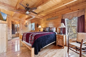 L’intérieur d’une cabane en bois, avec un lit, une chaise, une table de chevet, un ventilateur de plafond et une fenêtre avec des rideaux.
