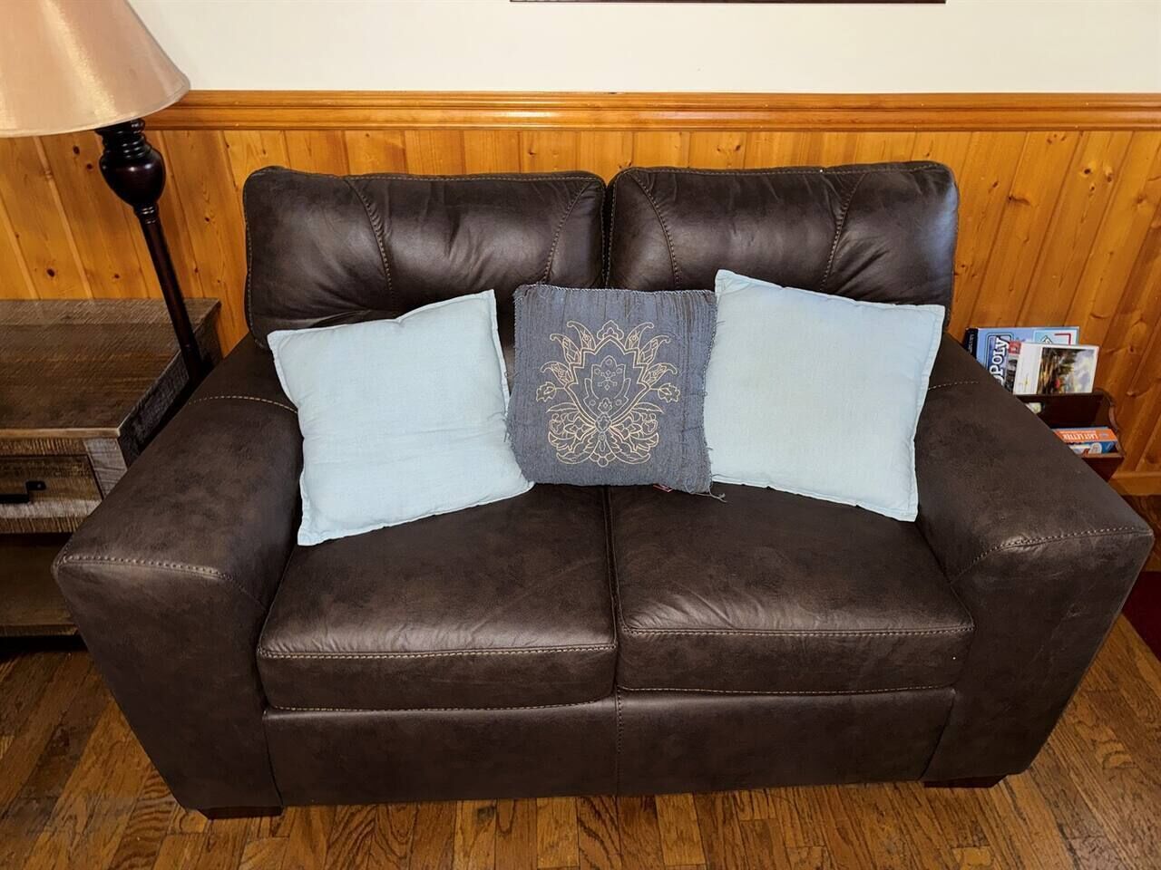 A brown leather sofa with two light blue pillows and a decorative pillow in a room with wooden paneling and a lamp.