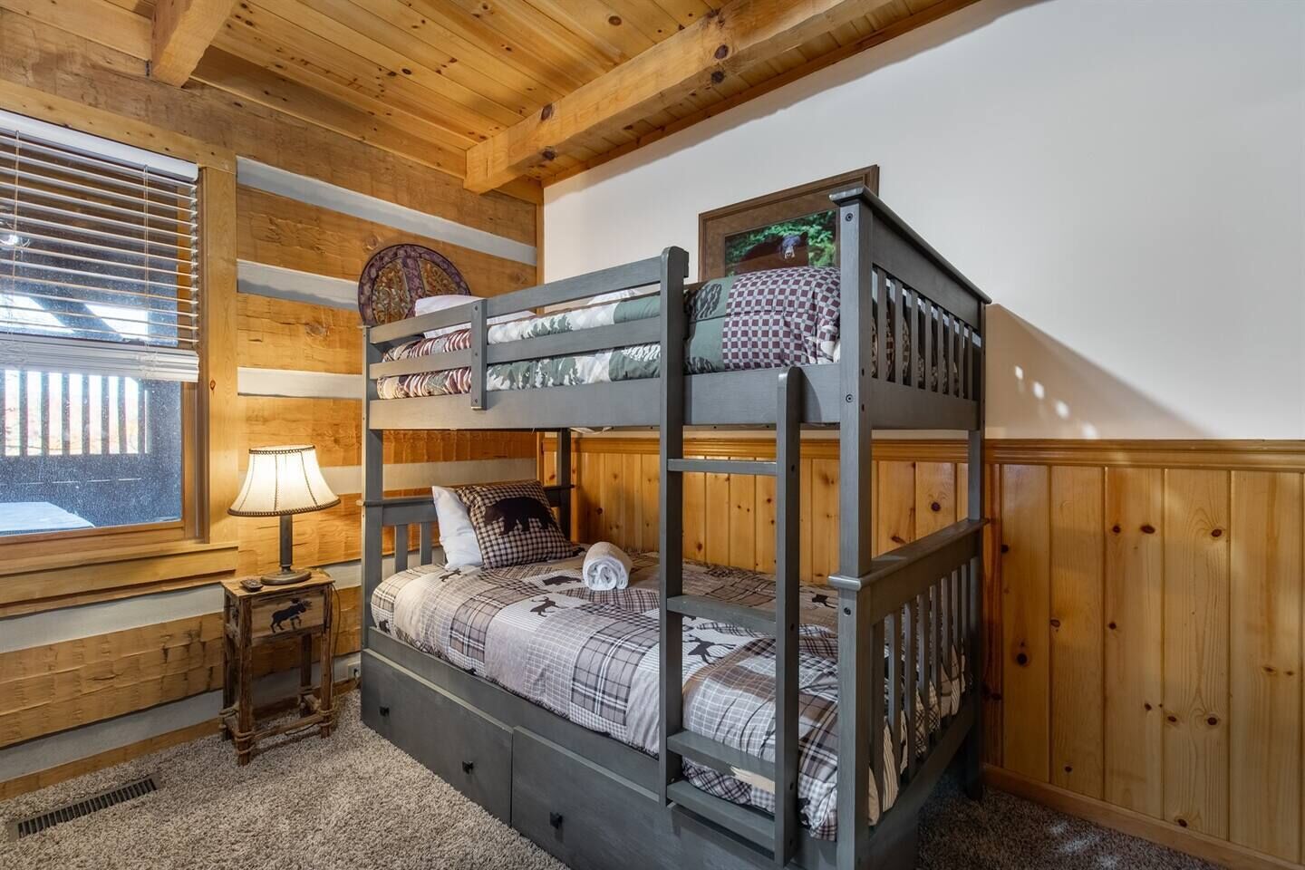 Une chambre avec un lit superposé, des murs en bois, une fenêtre avec des stores, une lampe et une petite table.