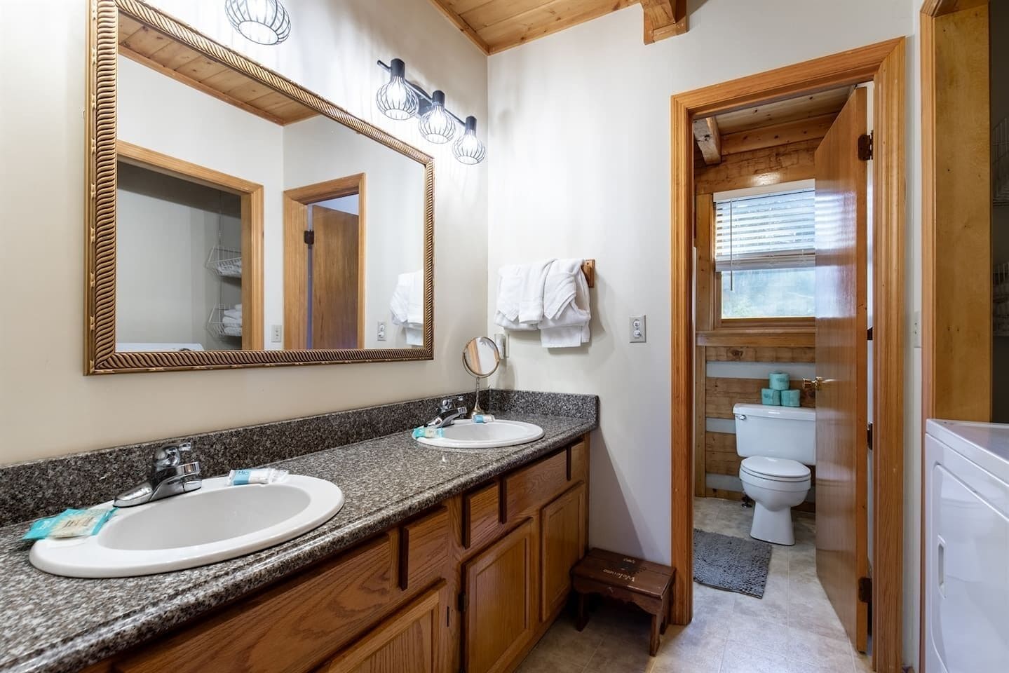 Un bagno con due lavandini, uno specchio grande, armadietti in legno e un WC visibile attraverso una porta aperta.