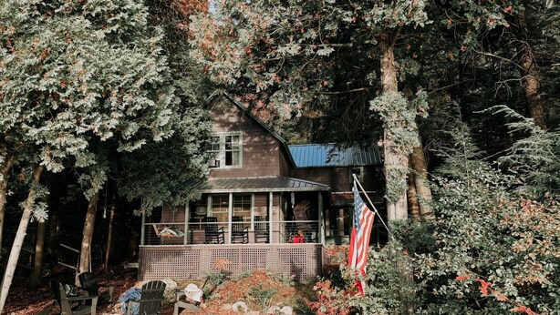 A cabin surrounded by dense forest, with a porch and outdoor seating area. - Cozy Waterfront Lake Cabin - New Listing - Adirondacks - Long Lost Lodge (Malone)