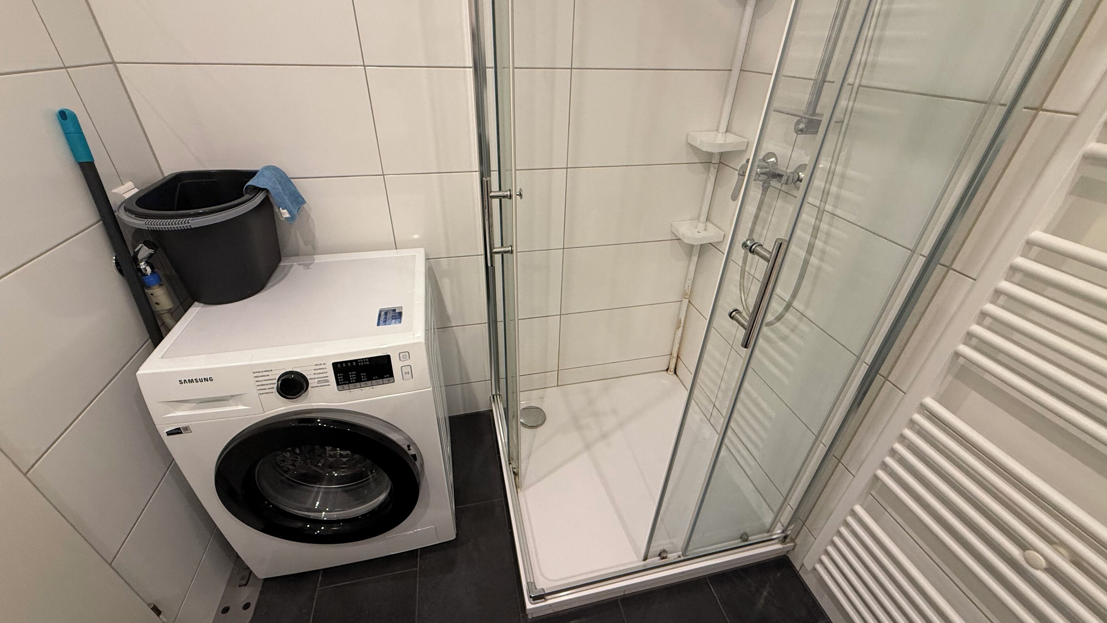 A compact bathroom with a washing machine, a glass shower enclosure, and a white tiled floor.
