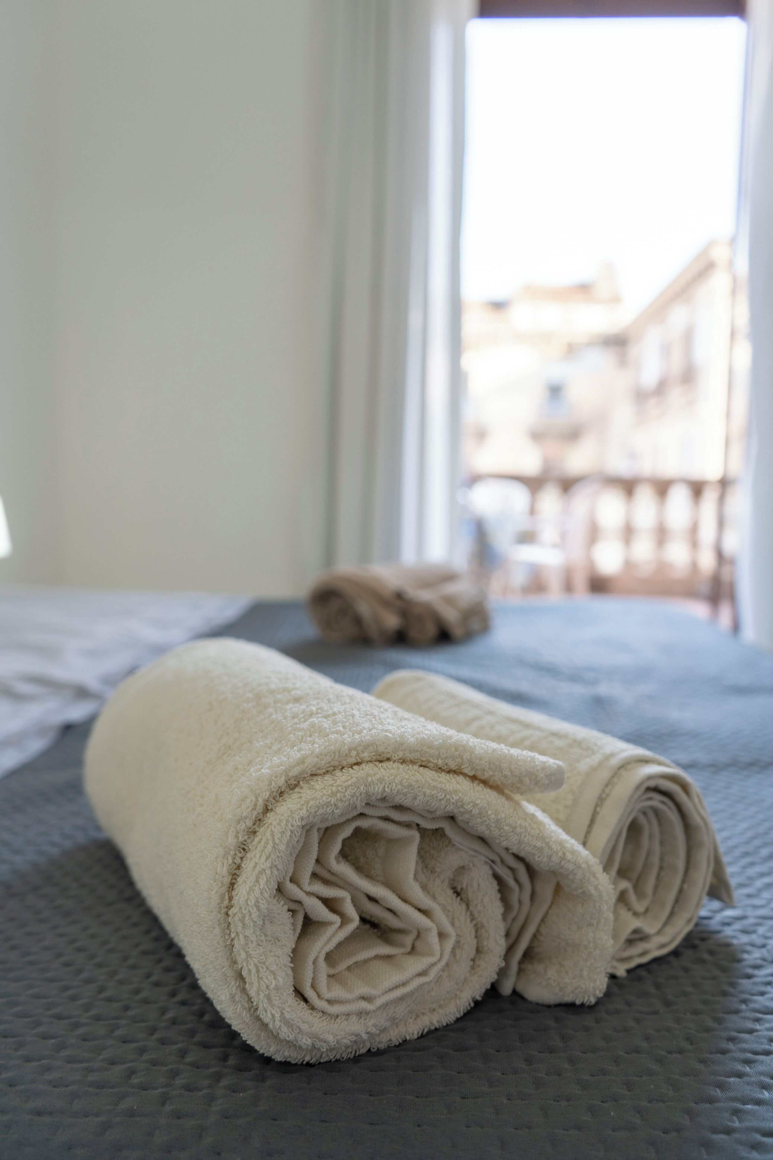 A neatly folded beige towel on a textured gray bedspread in a hotel room with a view of buildings through a window.