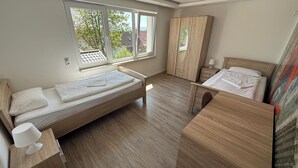 A small, well-lit room with two single beds, a wooden wardrobe, and a window providing a view of the outside.