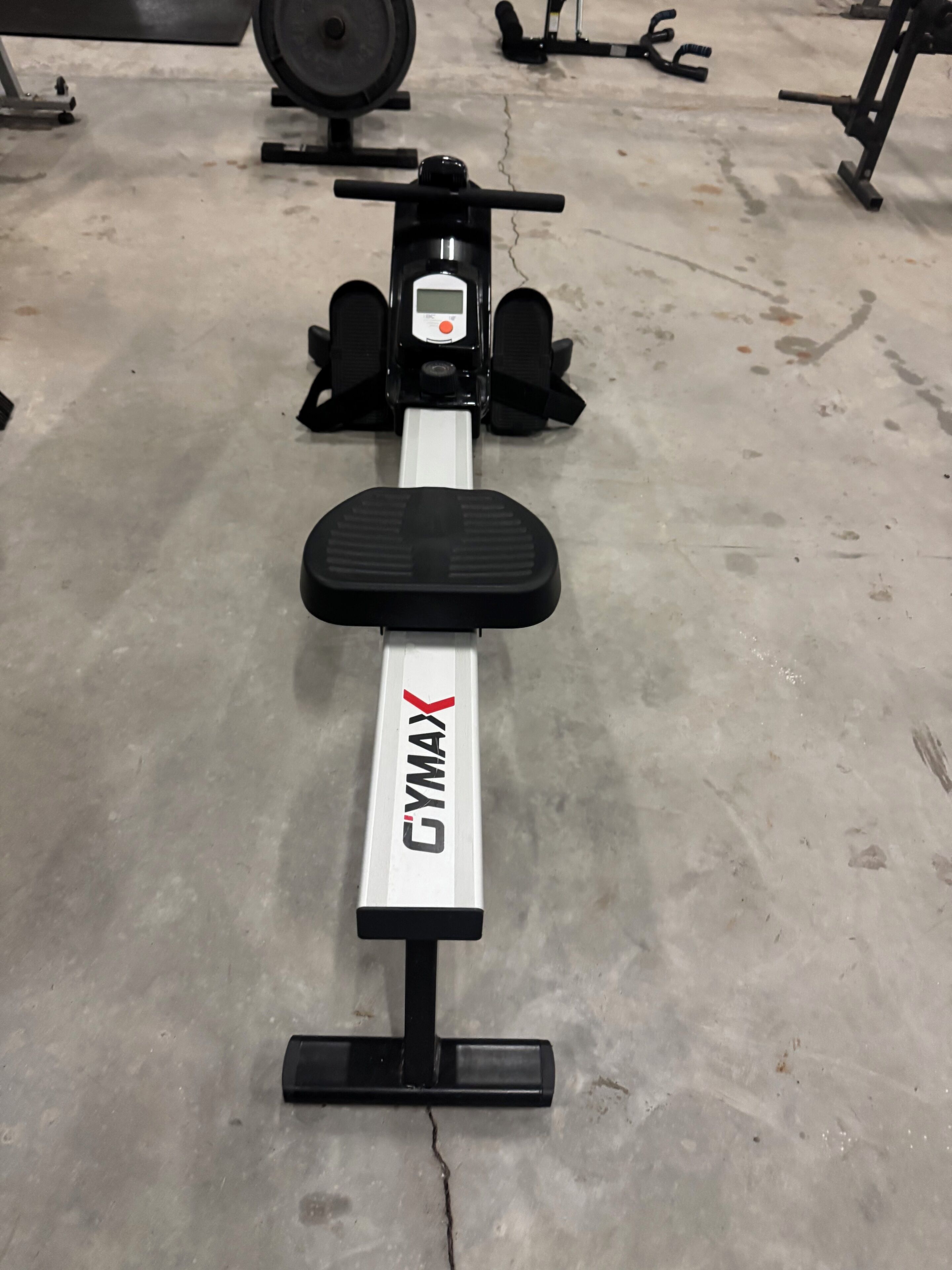 Ein Fitnessraum mit einer Bank mit der Aufschrift 'GYMAX' und verschiedenen Trainingsgeräten im Hintergrund.