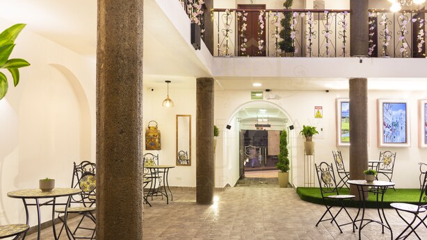 Un vestíbulo de hotel con una columna central, varias mesas y sillas, una escalera y un balcón con flores decorativas.